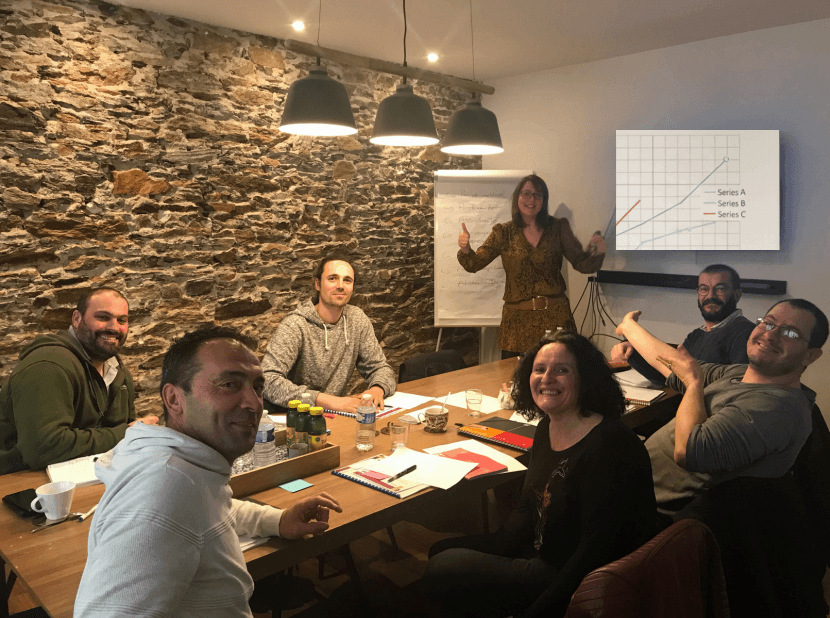 Atelier d’équipe autour d’une table : six participants souriants et l’animatrice debout devant un paperboard