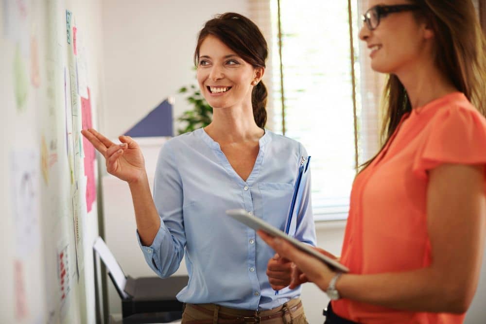 Deux femmes souriantes échangent debout devant un tableau couvert de notes adhésives, l’une tient une tablette