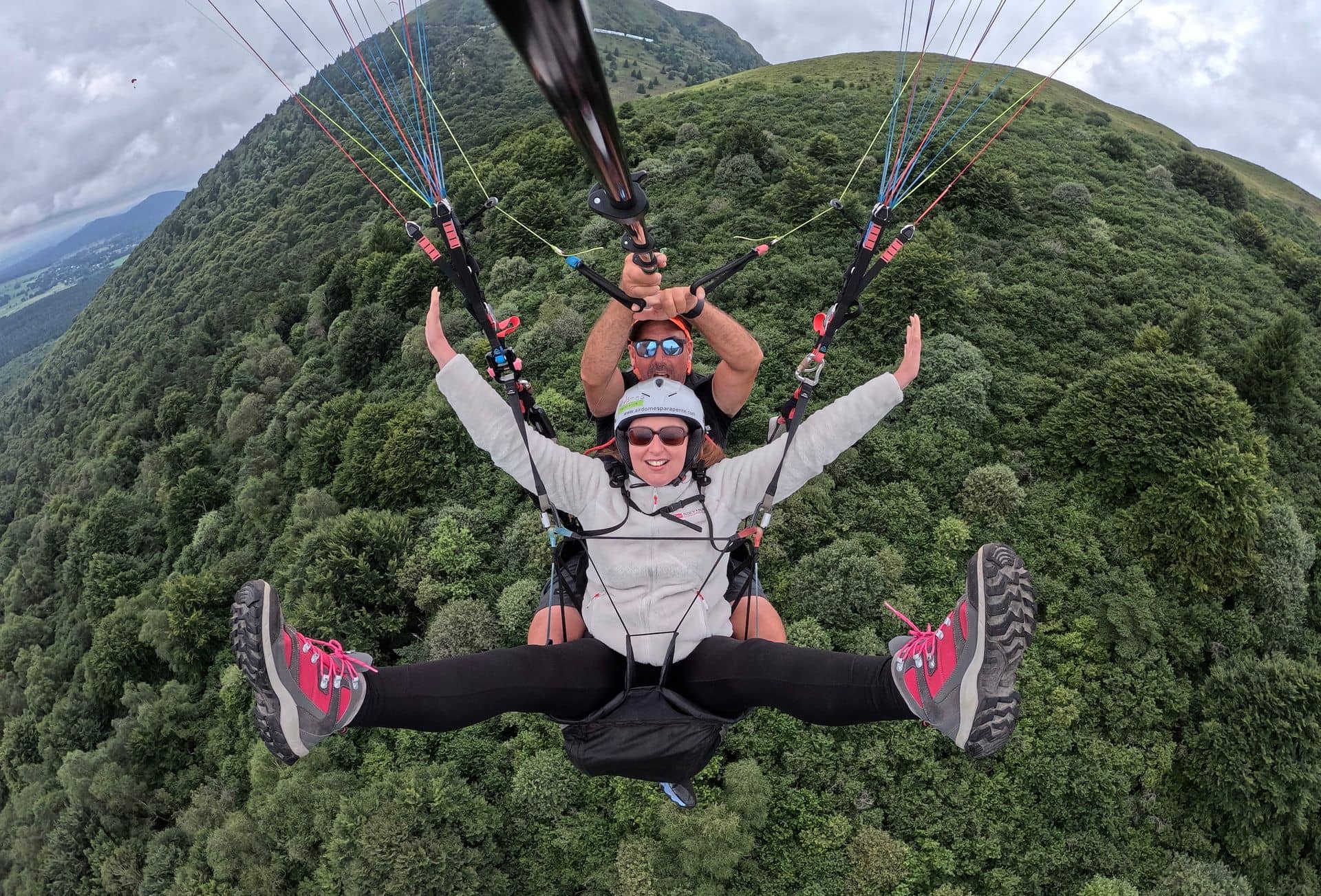 Nathalie Chiffoleau en vol en parapente biplace, bras ouverts et souriante, au-dessus d’une forêt