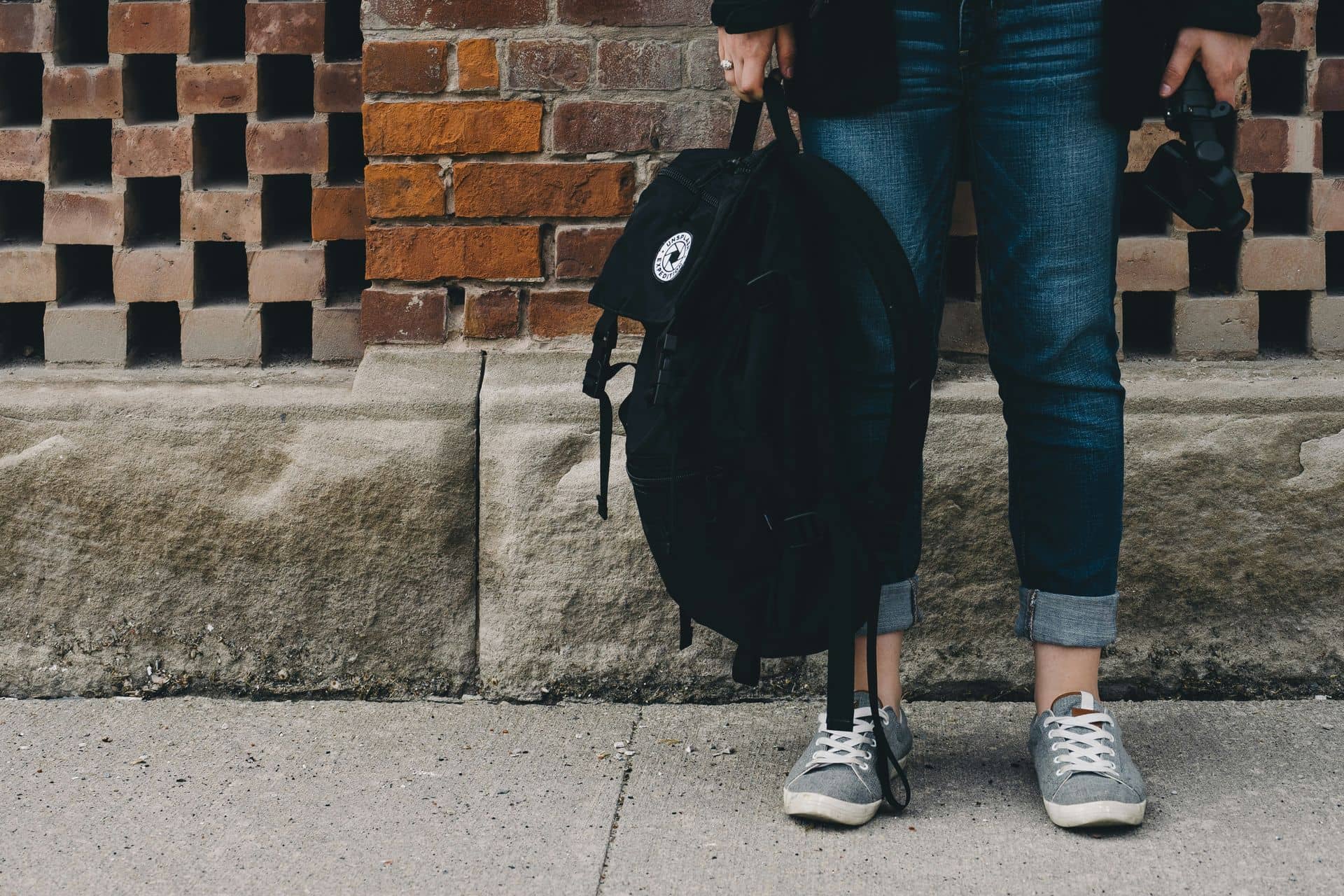 Jeune personne en jean et baskets, vue des jambes, tenant un sac à dos noir devant un mur de briques