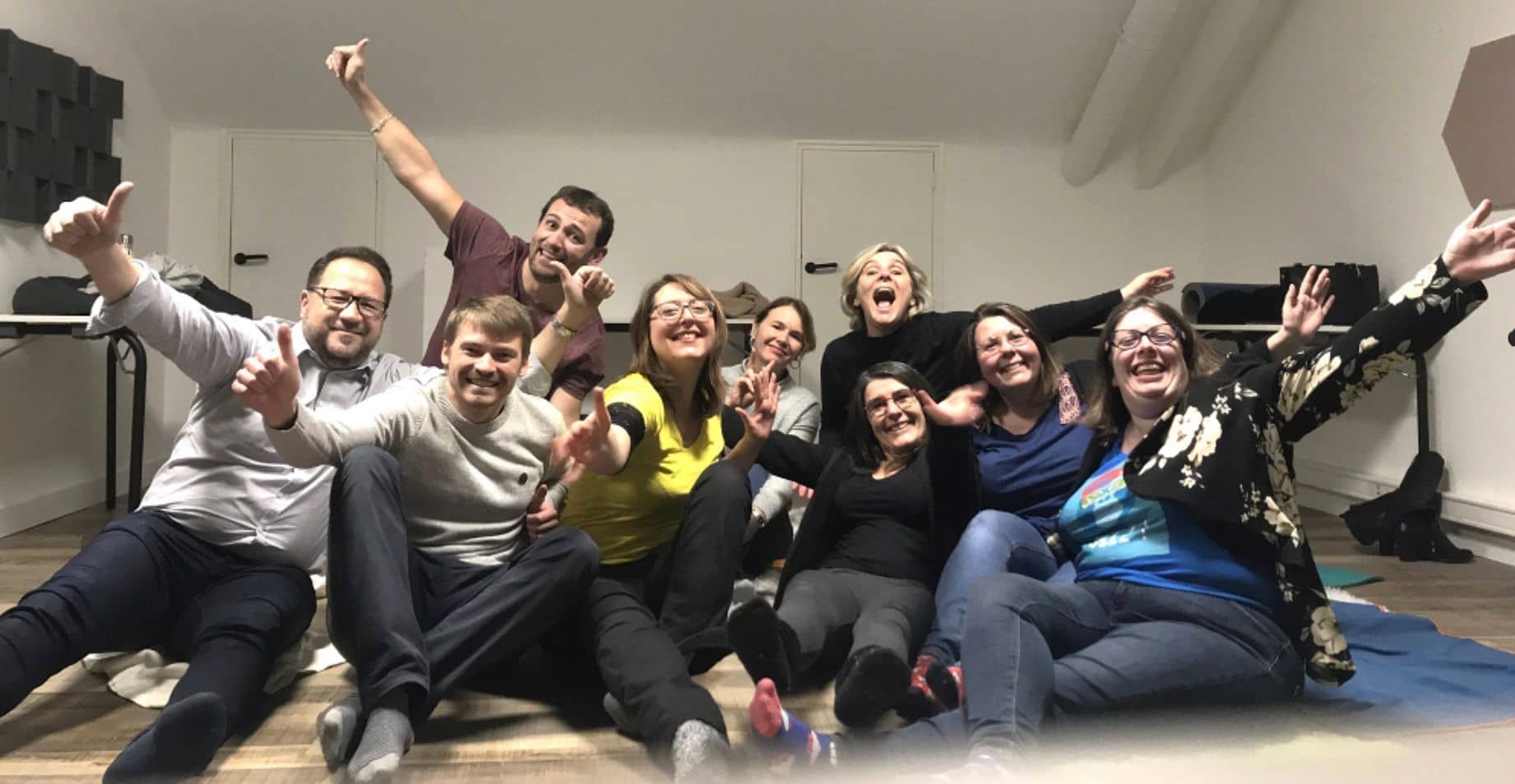 Groupe de neuf participants assis au sol, hilares, bras levés et pouces en l’air après une séance